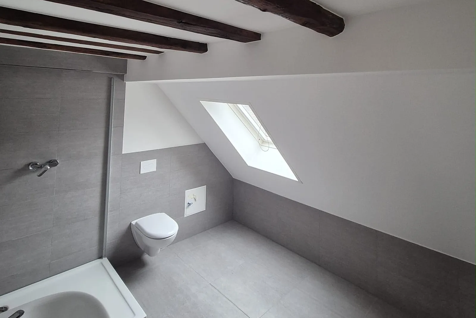 Bad unterm Dach mit Dachfenster, dunklen Deckenbalken und grauen Fliesen